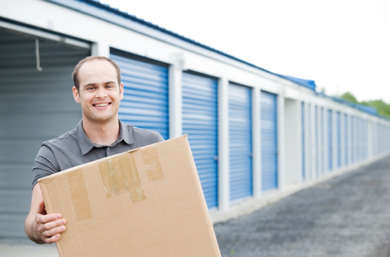 Smiling man holding box standing in front of storage units.