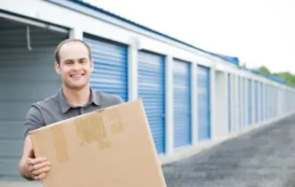 Smiling man holding box standing in front of storage units.
